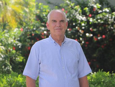 Portrait of Bruce McCord wearing a short-sleeved blue pinstriped shirt, standing outdoors in front of lush green foliage.