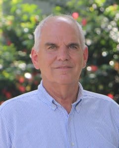 Portrait of Bruce McCord wearing a short-sleeved blue pinstriped shirt, standing outdoors in front of lush green foliage.