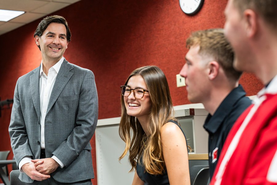 Instructor Brad Chandler stands smiling in a classroom alongside seated students engaged in conversation.