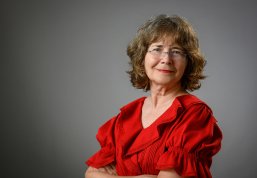 Portrait of a smiling woman with shoulder-length wavy hair and glasses, wearing a bright red ruffled top against a neutral gray background.