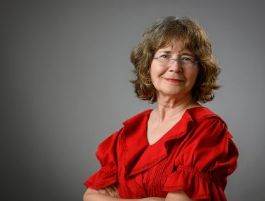 Portrait of a smiling woman with shoulder-length wavy hair and glasses, wearing a bright red ruffled top against a neutral gray background.