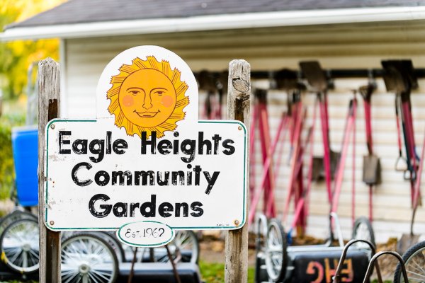 A sign reading 'Eagle Heights Community Gardens' topped with a smiling sun graphic, positioned in front of stored gardening tools and wheelbarrows.