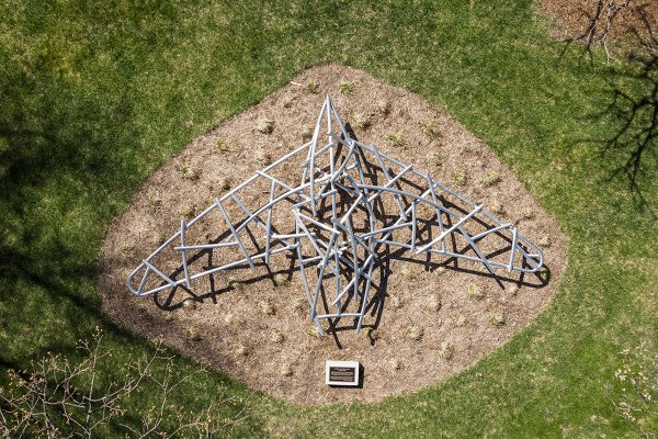An aerial view of a bird-shaped metal tube sculpture set within a mulch bed on a grassy lawn next to an informational plaque.