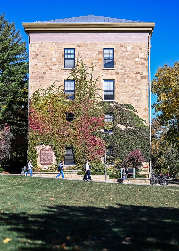 Pedestrians walk down Bascom Hill in front of North Hall -- a tall, stone building covered in red and green autumn ivy.