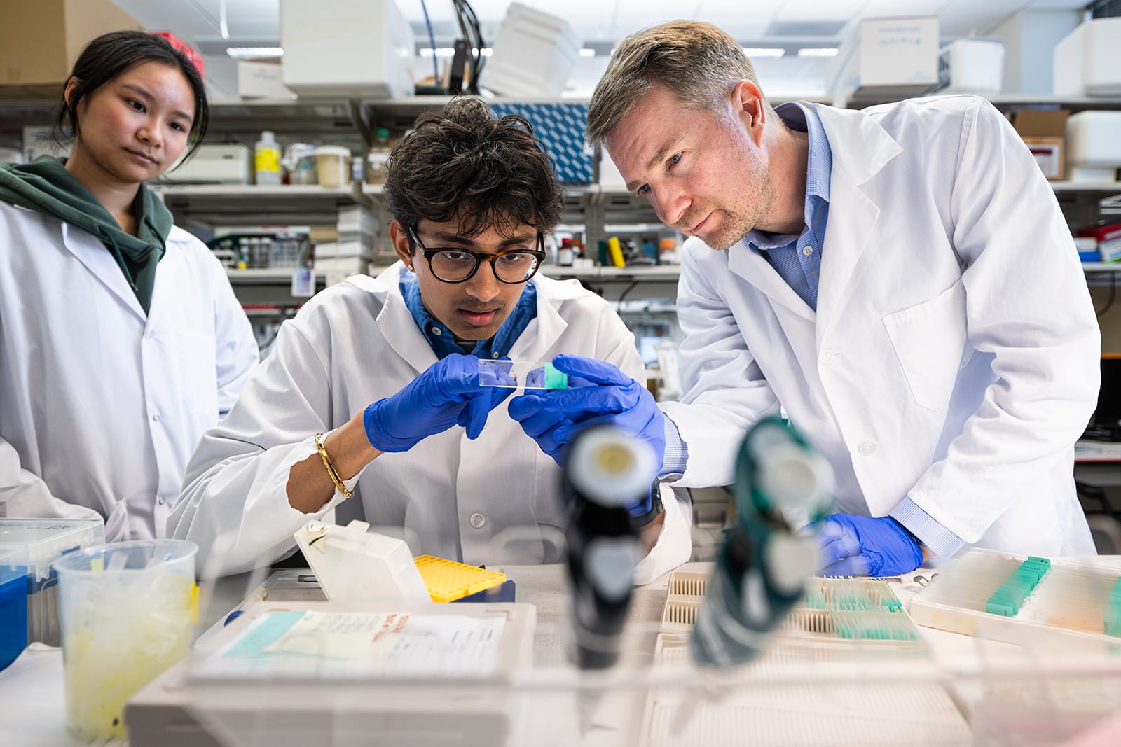 A scientist in a white lab coat guides a young researcher closely examining a glass sample slide at a laboratory workbench while a colleague observes from the background.