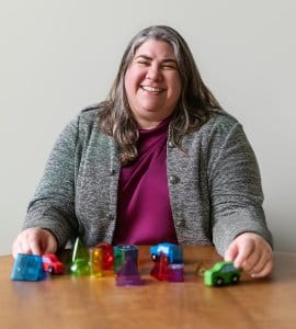 A smiling woman in a gray jacket sits at a wooden table with her hands positioned around small toy cars and colorful geometric blocks.