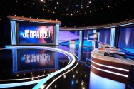 The empty studio set of the Jeopardy! game show features a large illuminated screen displaying the logo, a central host lectern, and contestant podiums under blue and purple studio lighting.