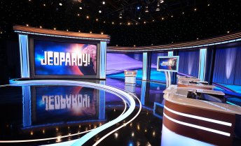 The empty studio set of the Jeopardy! game show features a large illuminated screen displaying the logo, a central host lectern, and contestant podiums under blue and purple studio lighting.