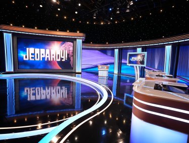 The empty studio set of the Jeopardy! game show features a large illuminated screen displaying the logo, a central host lectern, and contestant podiums under blue and purple studio lighting.