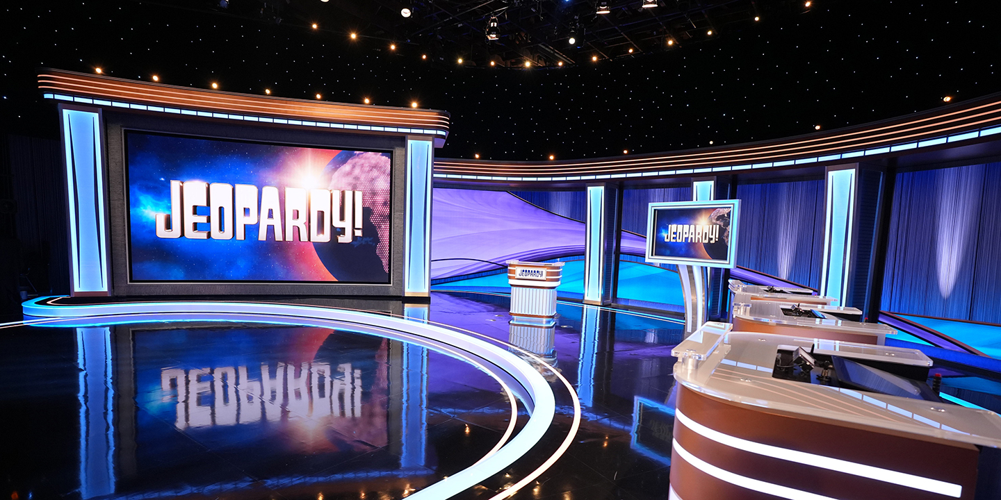 The empty studio set of the Jeopardy! game show features a large illuminated screen displaying the logo, a central host lectern, and contestant podiums under blue and purple studio lighting.