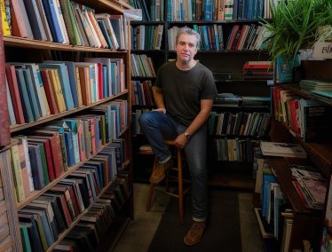 Jeff Oloizia sits on a wooden stool in a narrow aisle surrounded by floor-to-ceiling bookshelves filled with books.