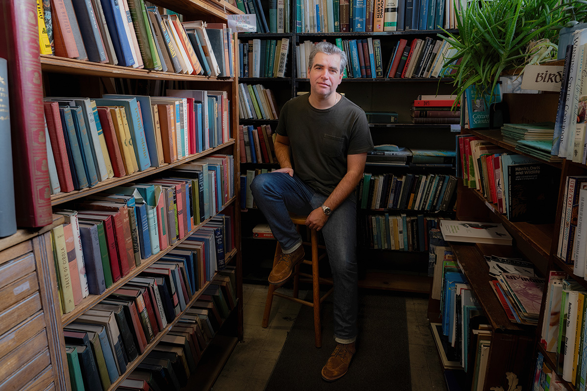 Jeff Oloizia sits on a wooden stool in a narrow aisle surrounded by floor-to-ceiling bookshelves filled with books.