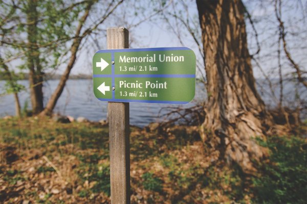 A wooden signpost on a shaded lakefront path showing directional arrows toward Memorial Union and Picnic Point.