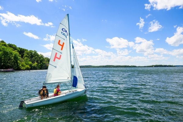 Two sailors guide a small sailboat across blue lake water under a partly cloudy sky.