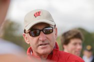 Close-up portrait of coach Mick Byrne wearing sunglasses and a beige Wisconsin logo cap outdoors.