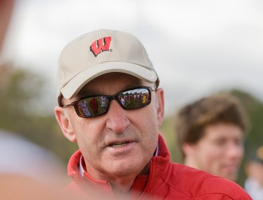Close-up portrait of coach Mick Byrne wearing sunglasses and a beige Wisconsin logo cap outdoors.