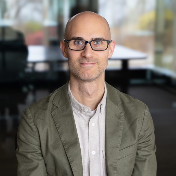 Portrait of Simon Goldberg wearing black-framed glasses and an olive green blazer.