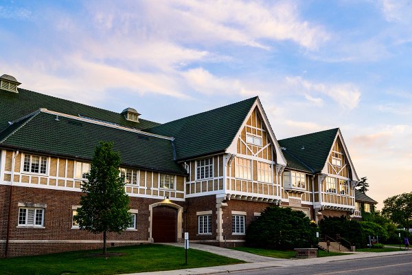 A large Tudor Revival building featuring a brick lower story and dark green tiled roofs stands beside a green lawn under a blue evening sky.
