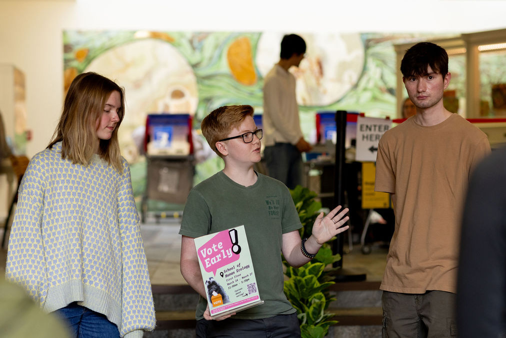 A student holding a 'Vote Early!' flyer gestures while speaking with two peers inside a campus voting location.