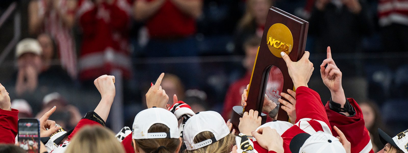 Members of Wisconsin's women's hockey team wearing red jerseys celebrate their victory by hoisting an NCAA championship trophy overhead.