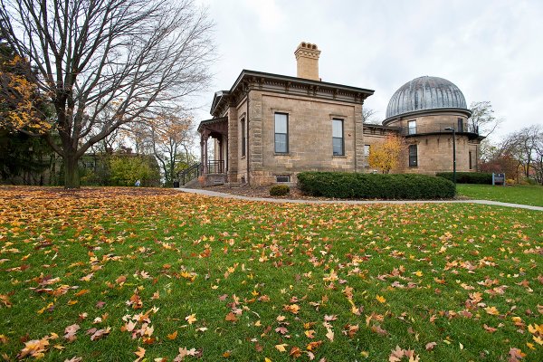 Fallen autumn leaves litter a green lawn in front of the historic stone Washburn Observatory building under an overcast sky.