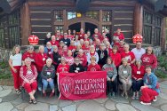 A large group of alumni wearing red and white Wisconsin apparel pose for a photo in front of a rustic log building while holding a red Wisconsin Alumni Association banner.