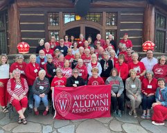 A large group of alumni wearing red and white Wisconsin apparel pose for a photo in front of a rustic log building while holding a red Wisconsin Alumni Association banner.