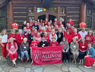 A large group of alumni wearing red and white Wisconsin apparel pose for a photo in front of a rustic log building while holding a red Wisconsin Alumni Association banner.