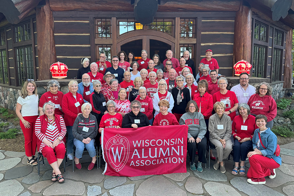 A large group of alumni wearing red and white Wisconsin apparel pose for a photo in front of a rustic log building while holding a red Wisconsin Alumni Association banner.