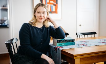Author Chloe Benjamin sitting at a wooden table resting her arm on a stack of copies of her novel Under Story.