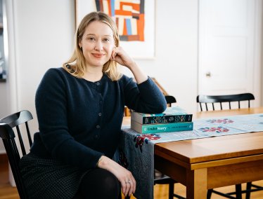 Author Chloe Benjamin sitting at a wooden table resting her arm on a stack of copies of her novel Under Story.