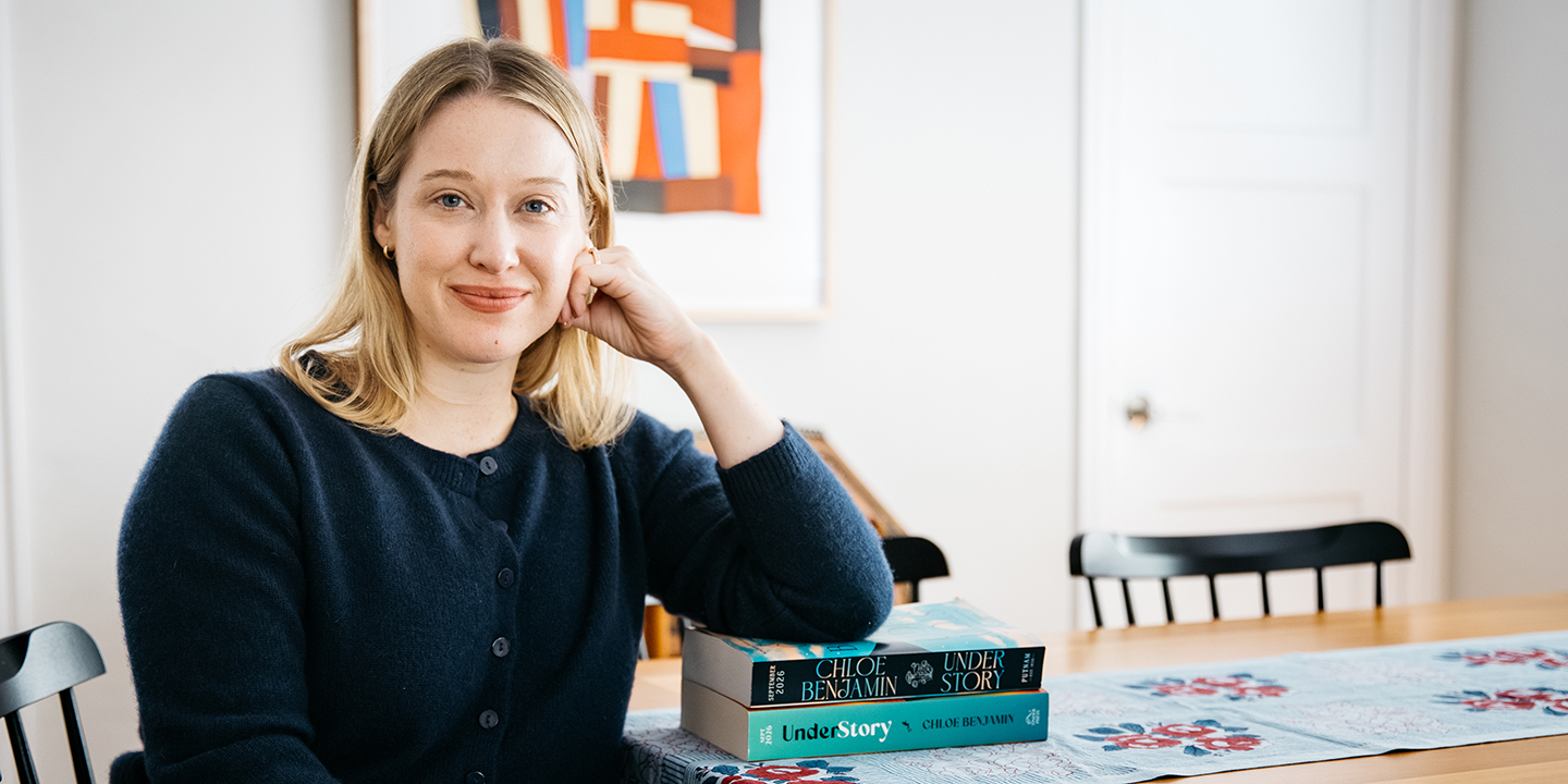 Author Chloe Benjamin sitting at a wooden table resting her arm on a stack of copies of her novel Under Story.