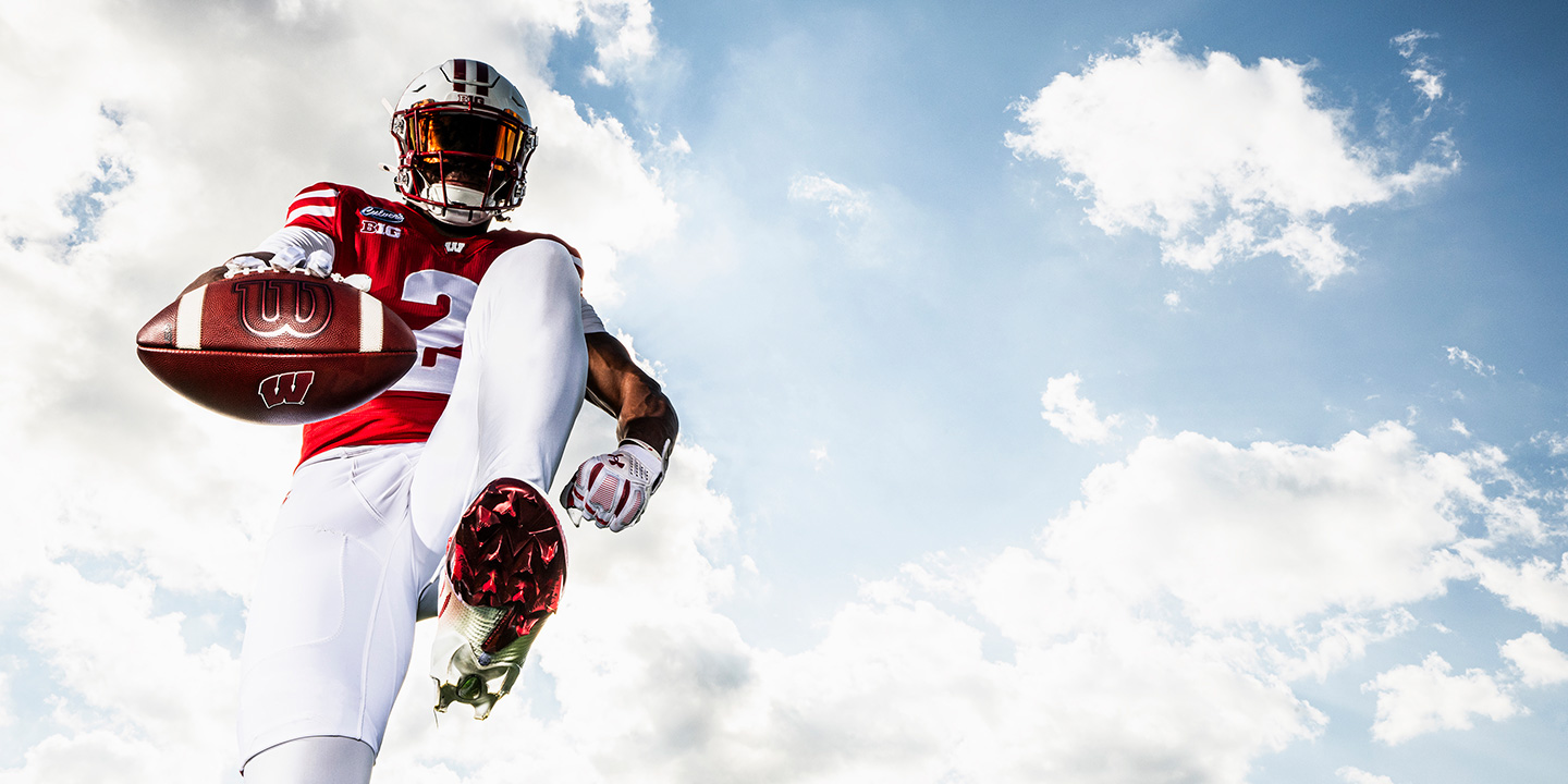 A low-angle shot of a Wisconsin Badgers football player high-stepping toward the camera and extending a football against a bright, cloudy blue sky.