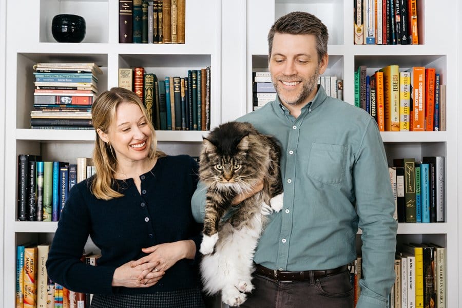 Author Chloe Benjamin and husband Nathan Jandl smile as they hold a large, fluffy cat in front of white bookshelves filled with books.