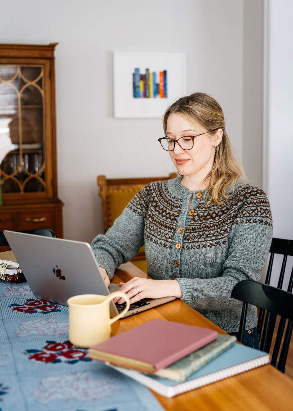 Chloe Benjamin wearing glasses and a patterned knit sweater works on a laptop at a dining table with notebooks and a mug nearby.