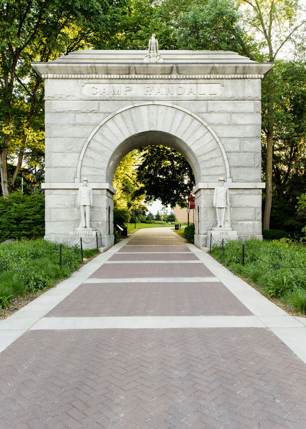 A historic stone archway labeled 'Camp Randall' flanked by two statues of Civil War soldiers, with a brick path leading through the arch into a tree-lined area.