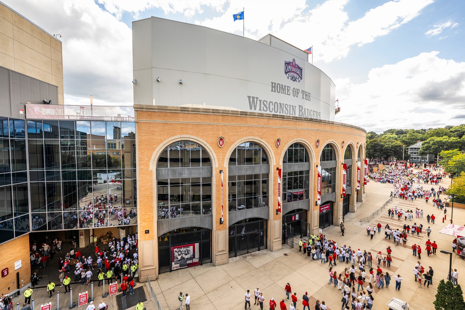 An exterior aerial view of Camp Randall Stadium featuring large brick entrance arches and crowds of fans walking along the plaza below.