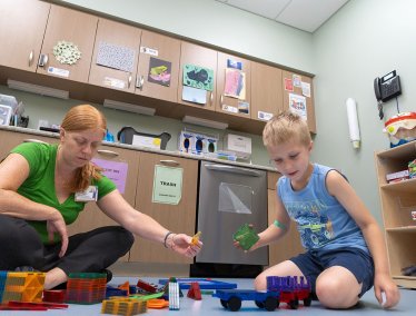 A woman in a green shirt sits on the floor playing with colorful magnetic building tiles alongside a young boy.