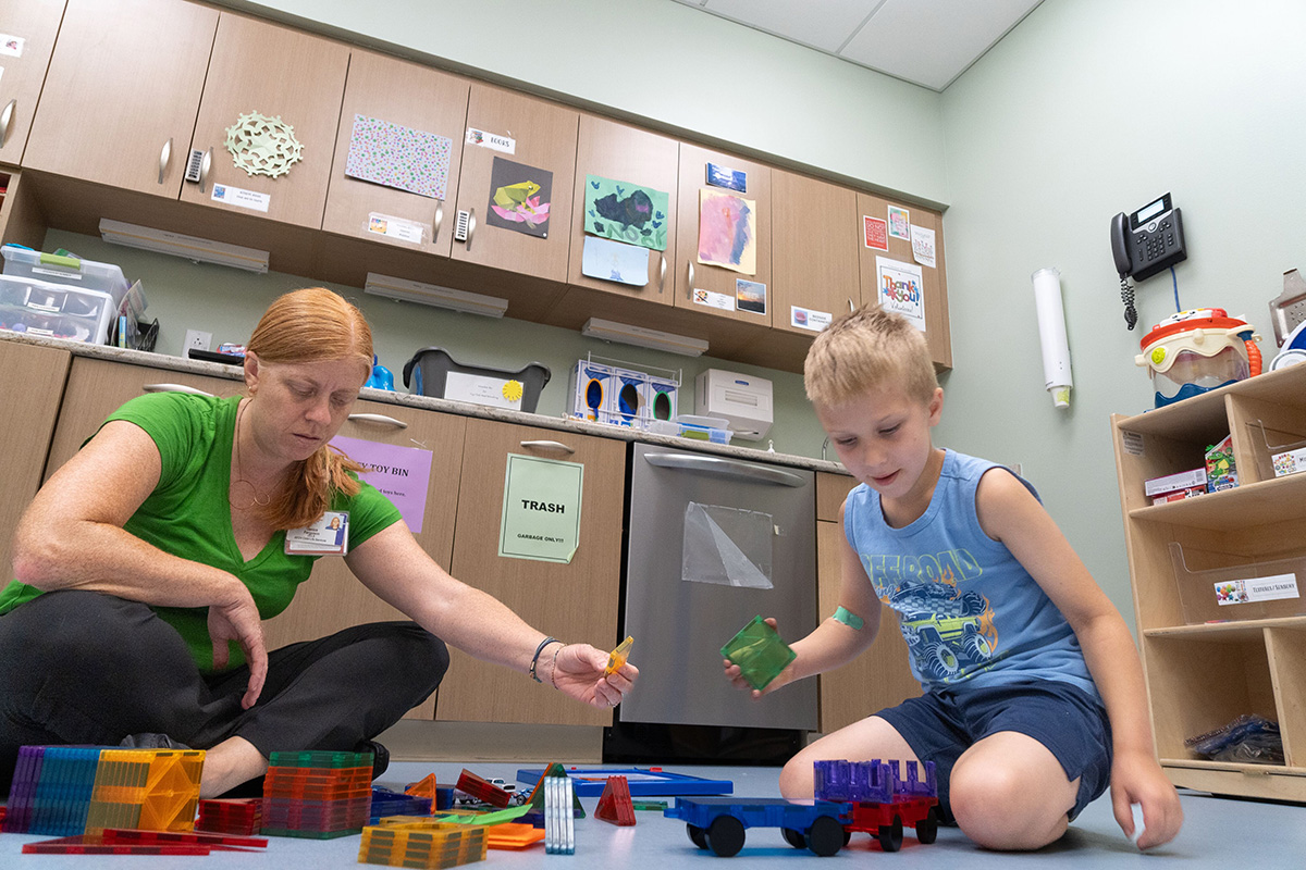 A woman in a green shirt sits on the floor playing with colorful magnetic building tiles alongside a young boy.