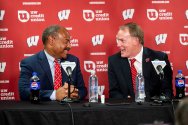 Eric Wilcots and Shawn Eichorst, in suits, smile and converse while seated at a press conference table in front of a red Wisconsin-branded backdrop.