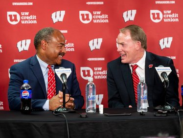 Eric Wilcots and Shawn Eichorst, in suits, smile and converse while seated at a press conference table in front of a red Wisconsin-branded backdrop.