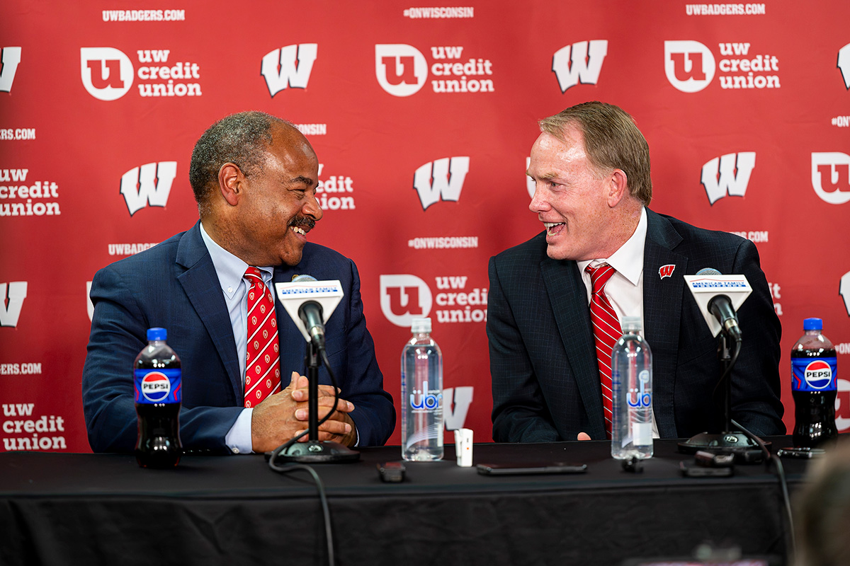 Eric Wilcots and Shawn Eichorst, in suits, smile and converse while seated at a press conference table in front of a red Wisconsin-branded backdrop.