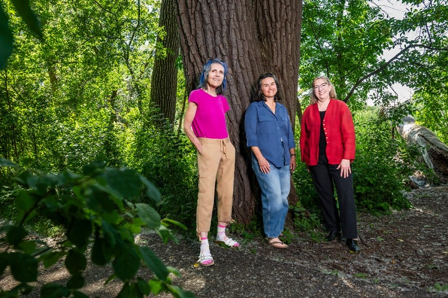 Three women stand together in front of a large tree trunk in a lush, wooded outdoor setting.