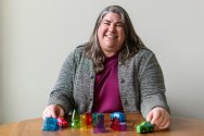 A smiling woman in a gray jacket sits at a wooden table with her hands positioned around small toy cars and colorful geometric blocks.