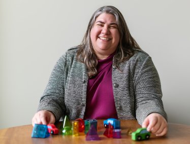 A smiling woman in a gray jacket sits at a wooden table with her hands positioned around small toy cars and colorful geometric blocks.
