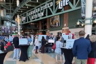 Attendees gather around display tables and presentation boards inside the spacious Lambeau Field Atrium during an event.