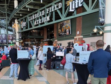Attendees gather around display tables and presentation boards inside the spacious Lambeau Field Atrium during an event.