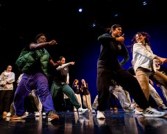 A group of dancers dressed in casual streetwear performs a high-energy dance routine on stage under blue lighting.