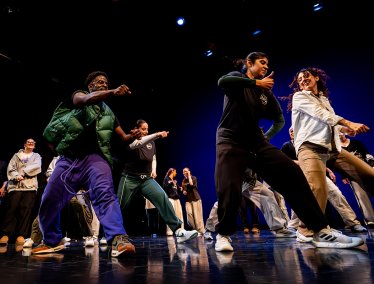 A group of dancers dressed in casual streetwear performs a high-energy dance routine on stage under blue lighting.