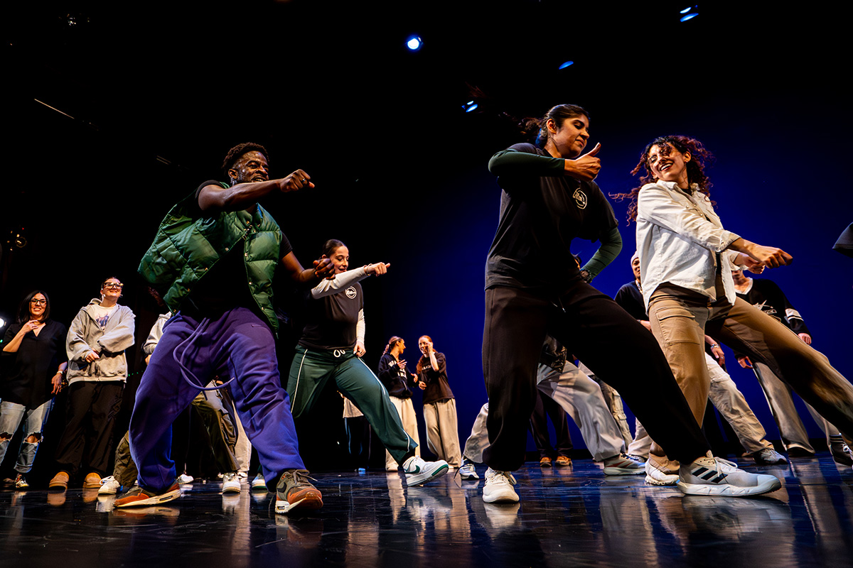 A group of dancers dressed in casual streetwear performs a high-energy dance routine on stage under blue lighting.